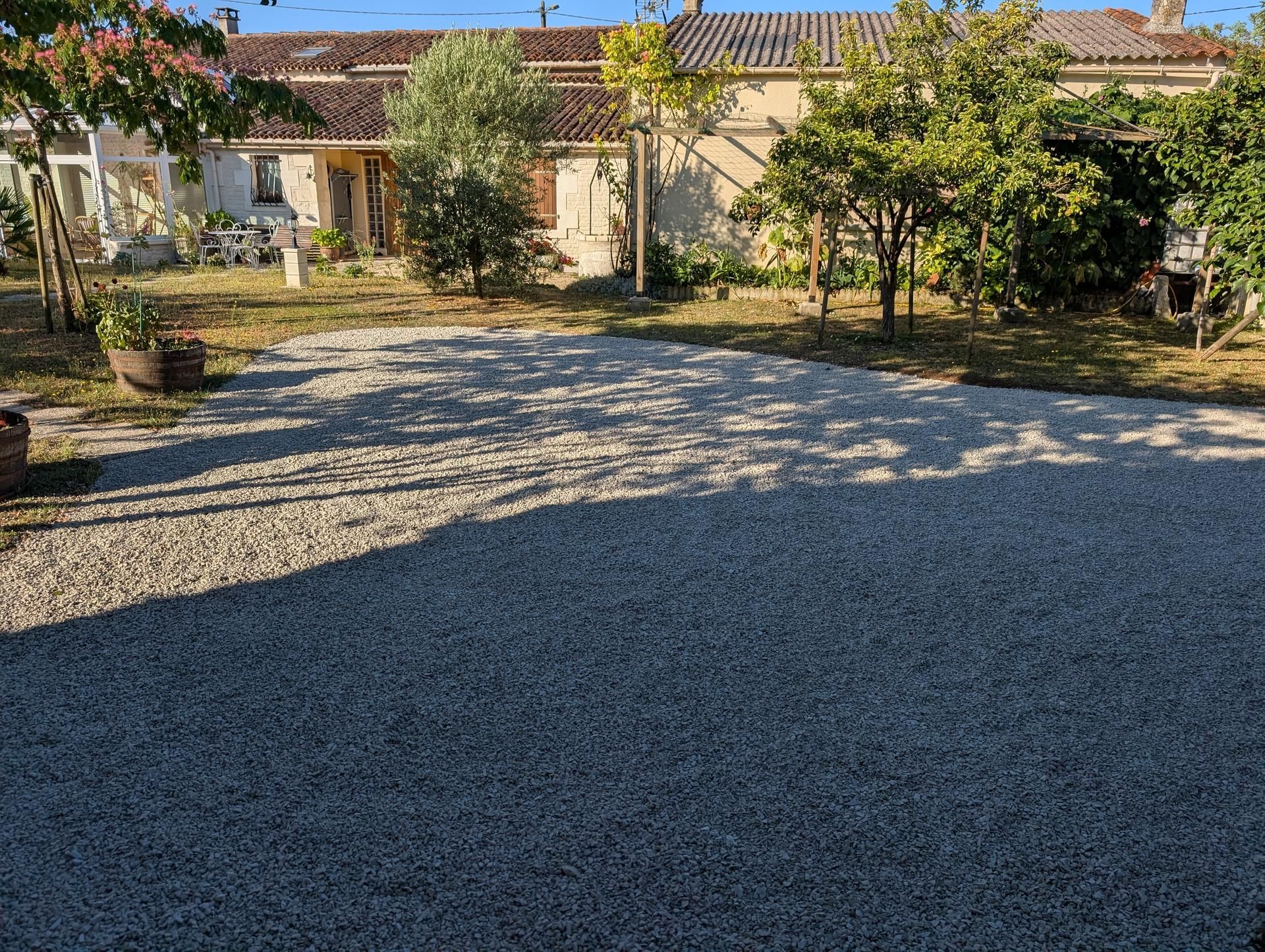 Allée de gravier devant une maison, arbres sur le côté, journée ensoleillée.