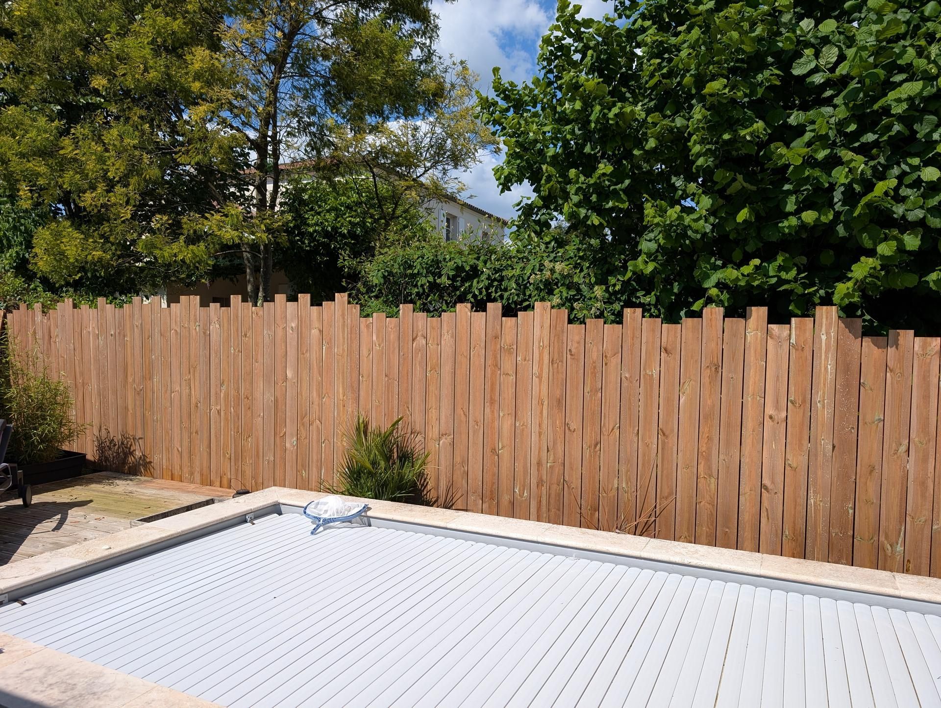 Clôture en bois bordant une piscine, arbres en arrière-plan, journée ensoleillée.
