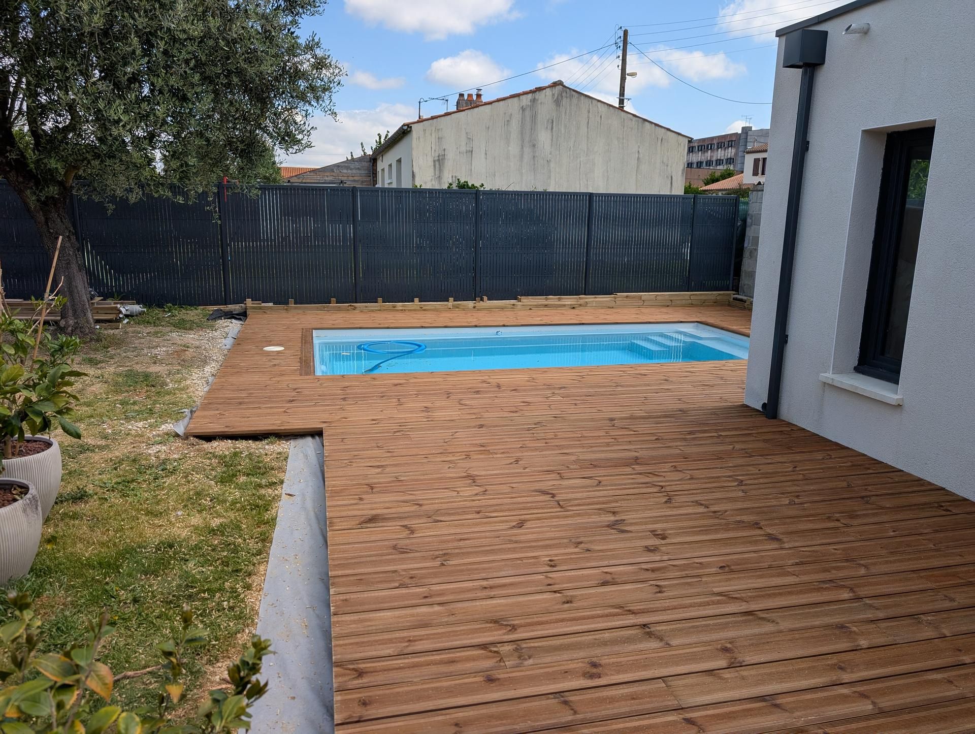 Une terrasse en bois entoure une piscine rectangulaire dans un jardin, à côté d'une maison blanche, avec une clôture sombre.