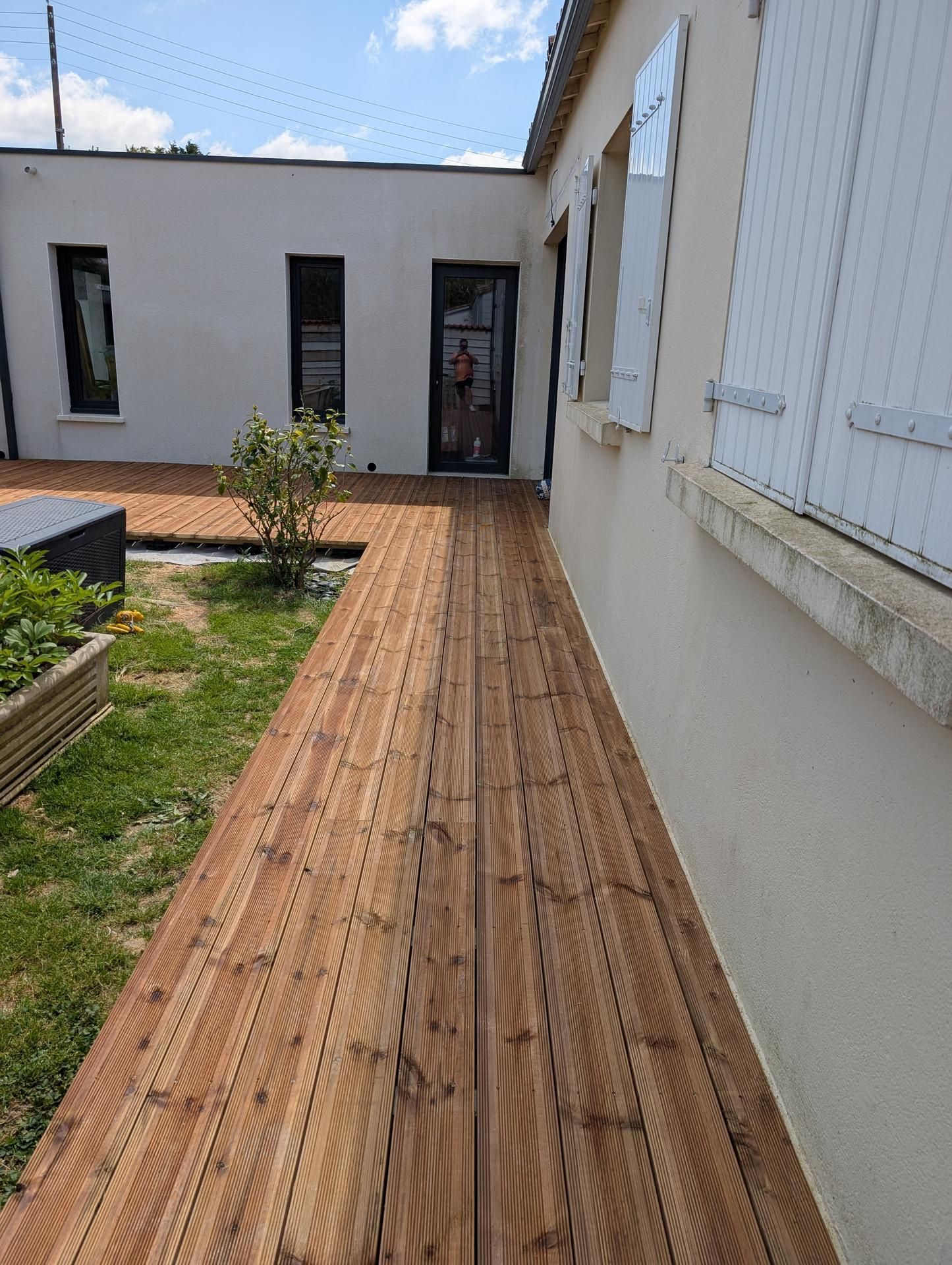 Allée en bois longeant un bâtiment clair aux fenêtres à volets ; petit jardin.
