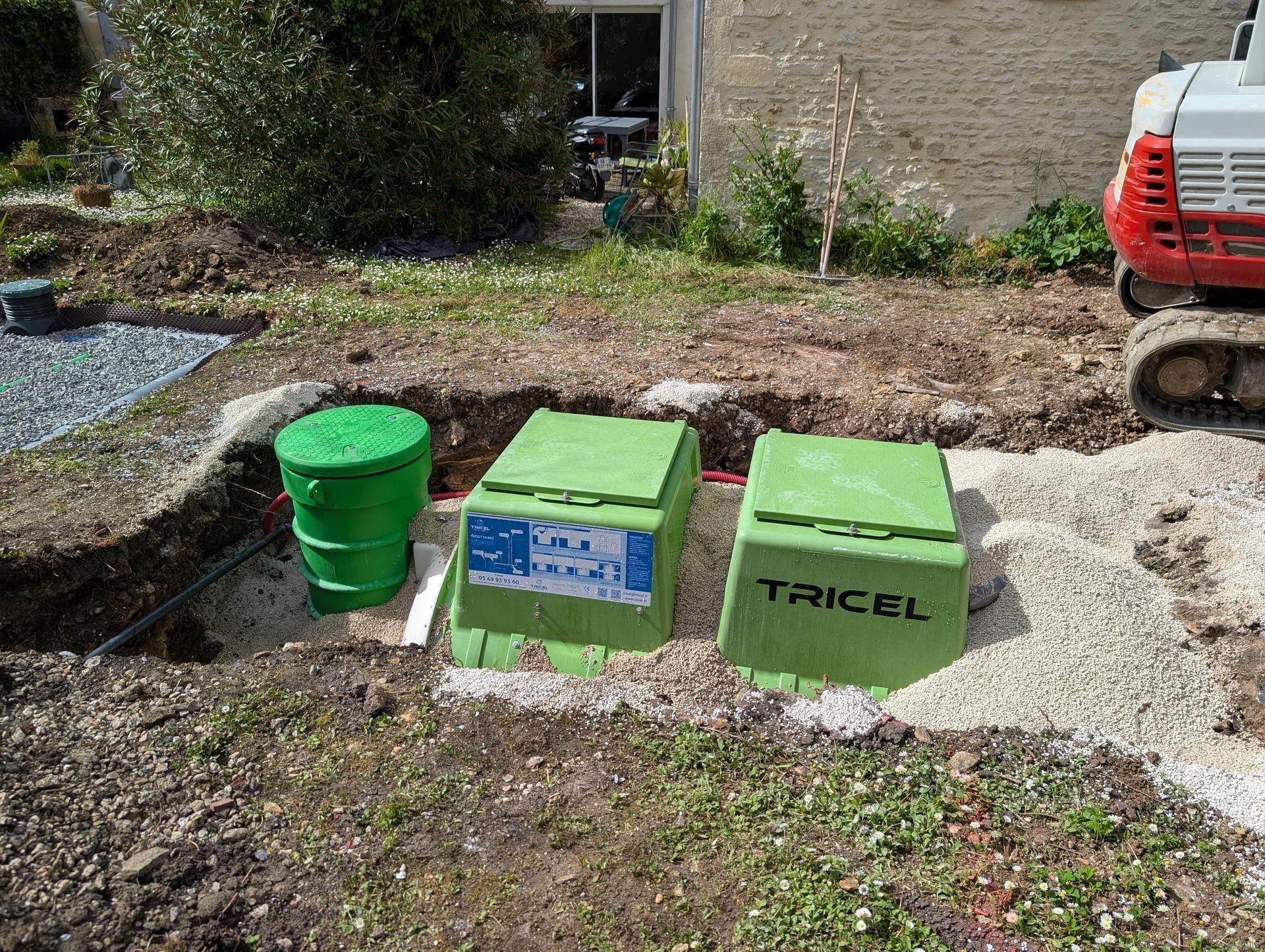 Des fosses septiques vertes sont installées dans une fosse à terre à l'extérieur d'un bâtiment, une petite excavatrice étant visible.