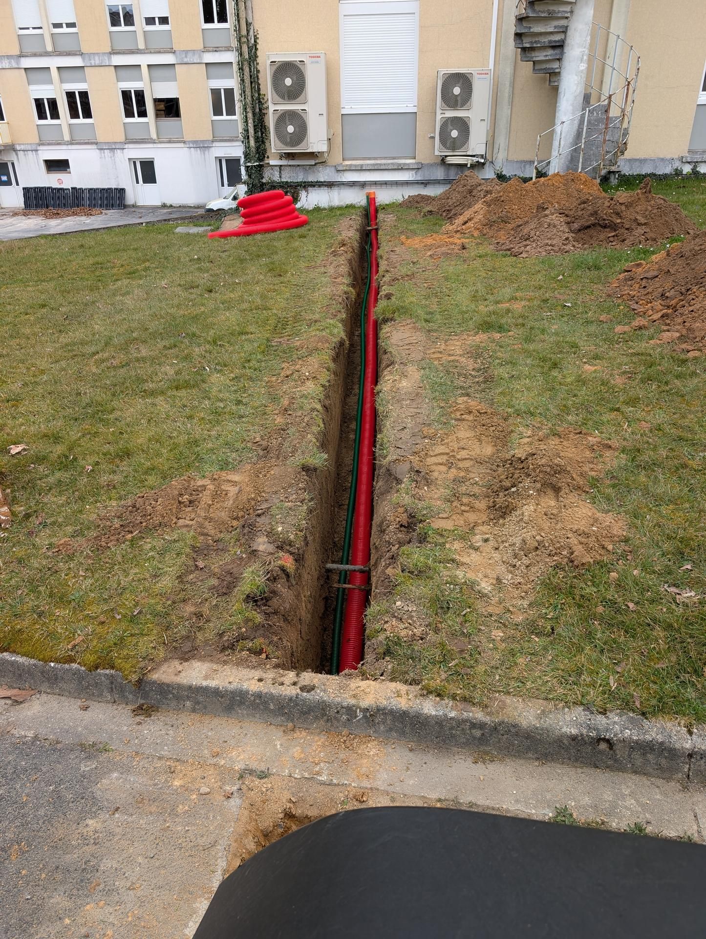 Un tuyau rouge en conduit se trouve dans une tranchée étroite creusée dans l'herbe, près d'un bâtiment et d'un trottoir.