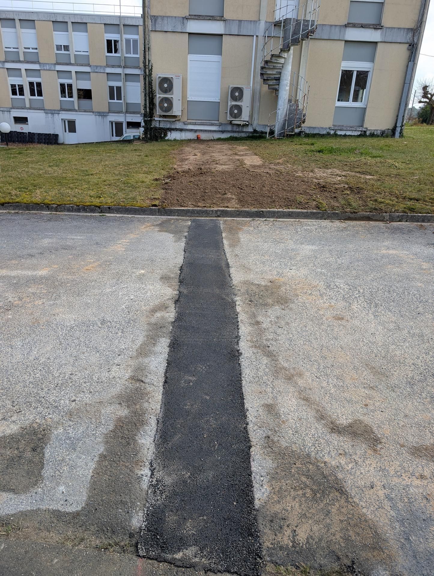 Réparation d'une bande d'asphalte noir traversant un stationnement en asphalte gris, menant à un bâtiment avec une zone herbeuse.