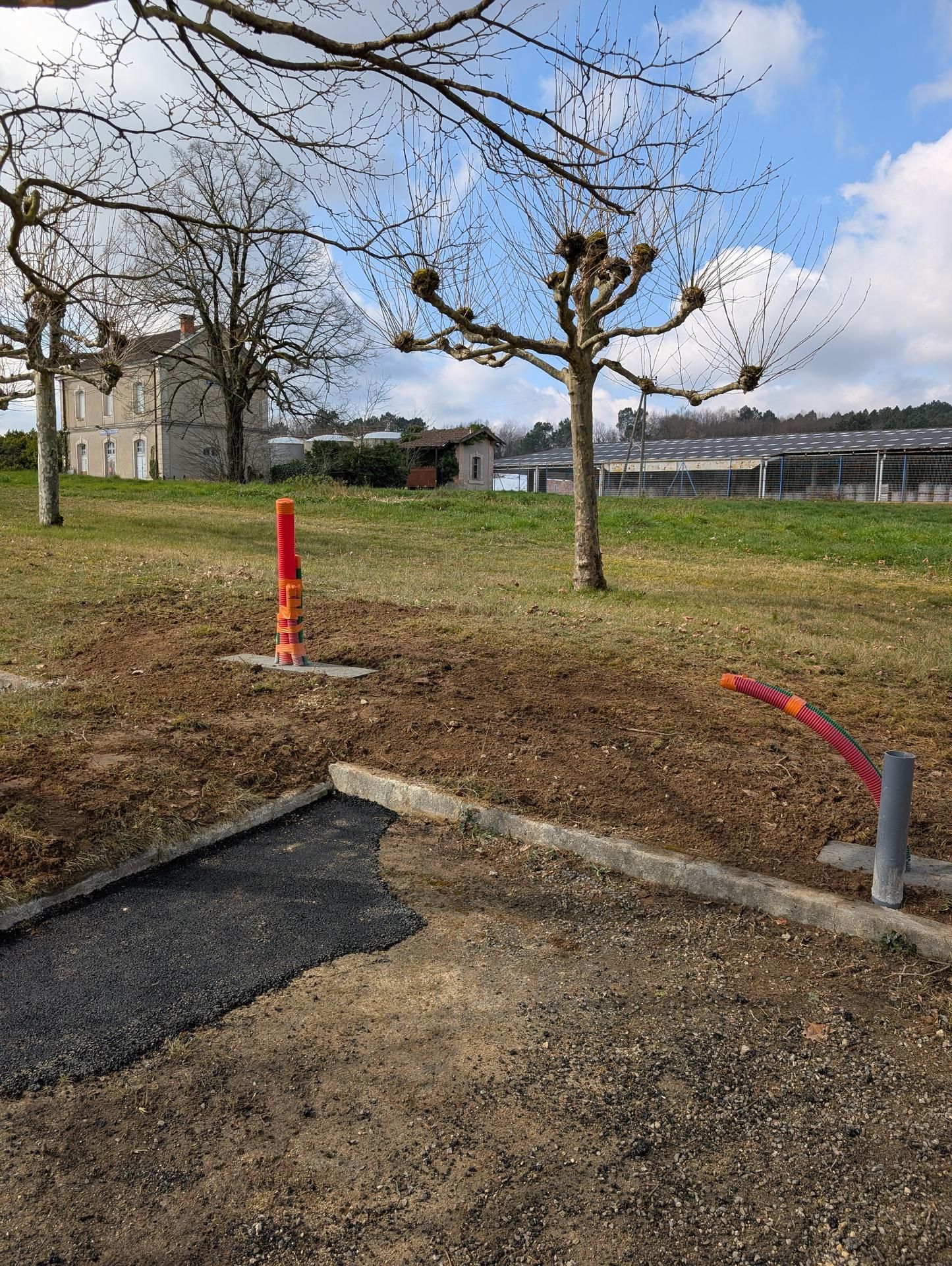 Parc avec des arbres dénudés, des poteaux rouges et gris, et une allée partiellement pavée sous un ciel nuageux.