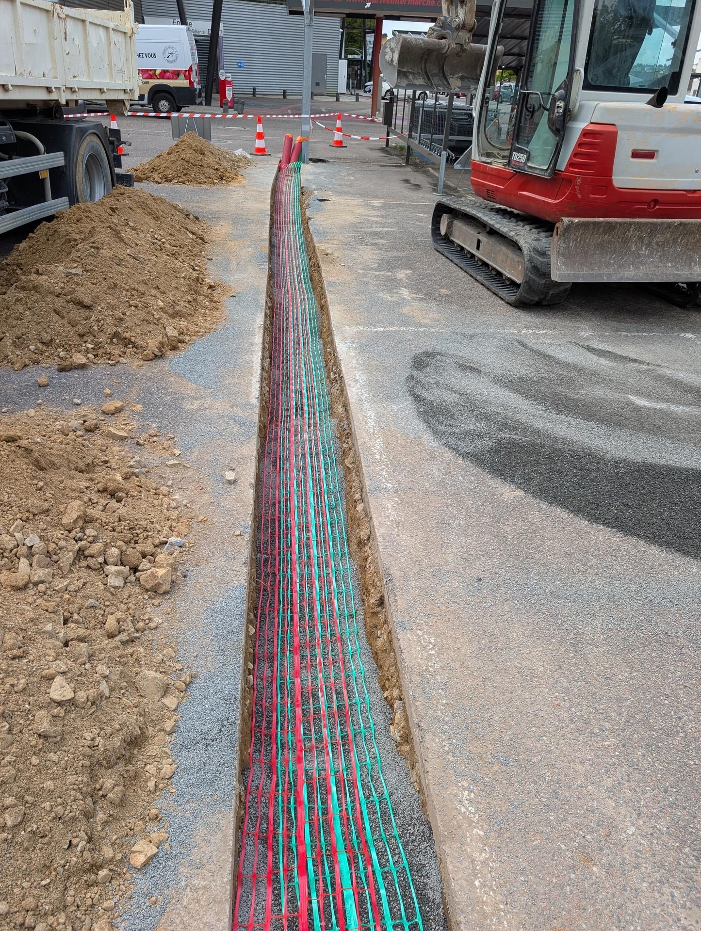 Tranchée avec câbles rouges et verts dans la chaussée ; camion, excavatrice et tas de terre visibles.