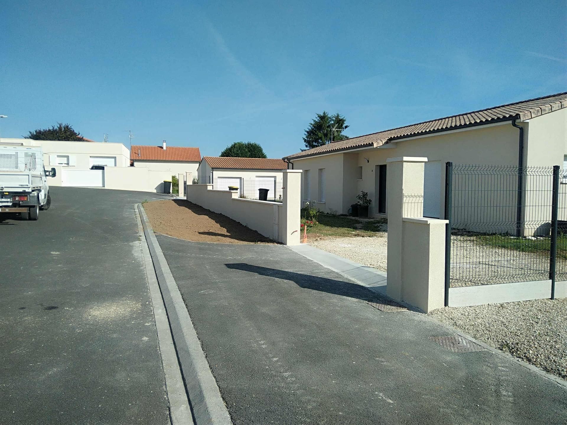 Une rue pavée bordée de maisons. Un muret entoure la maison de droite, qui possède un portail noir. Ciel bleu clair.