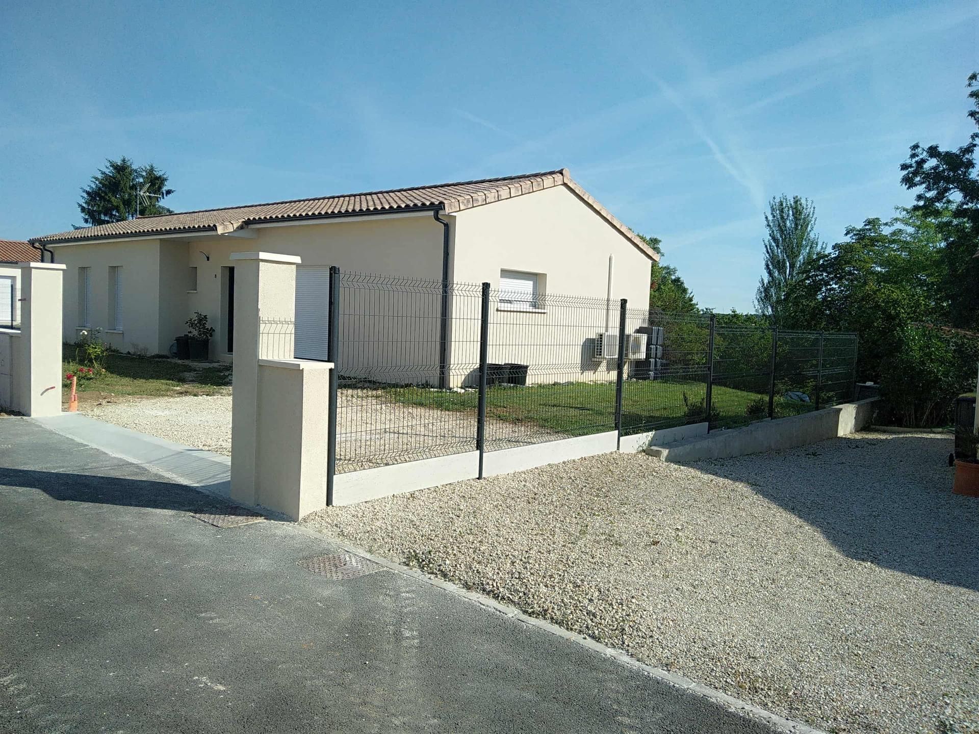 Maison beige avec une clôture et une allée de gravier sous un ciel d'un bleu éclatant.