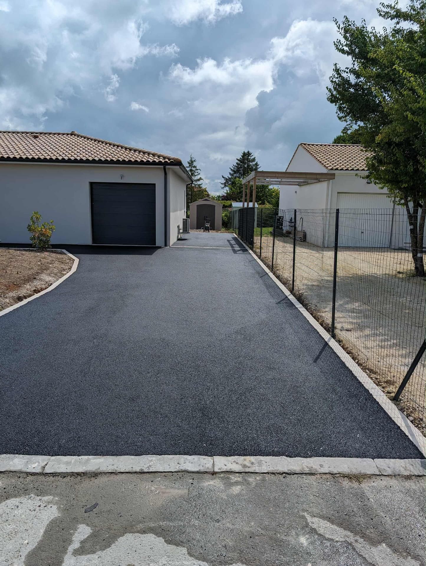 Allée asphaltée menant à des bâtiments blancs sous un ciel nuageux, bordée de béton et d'une clôture.