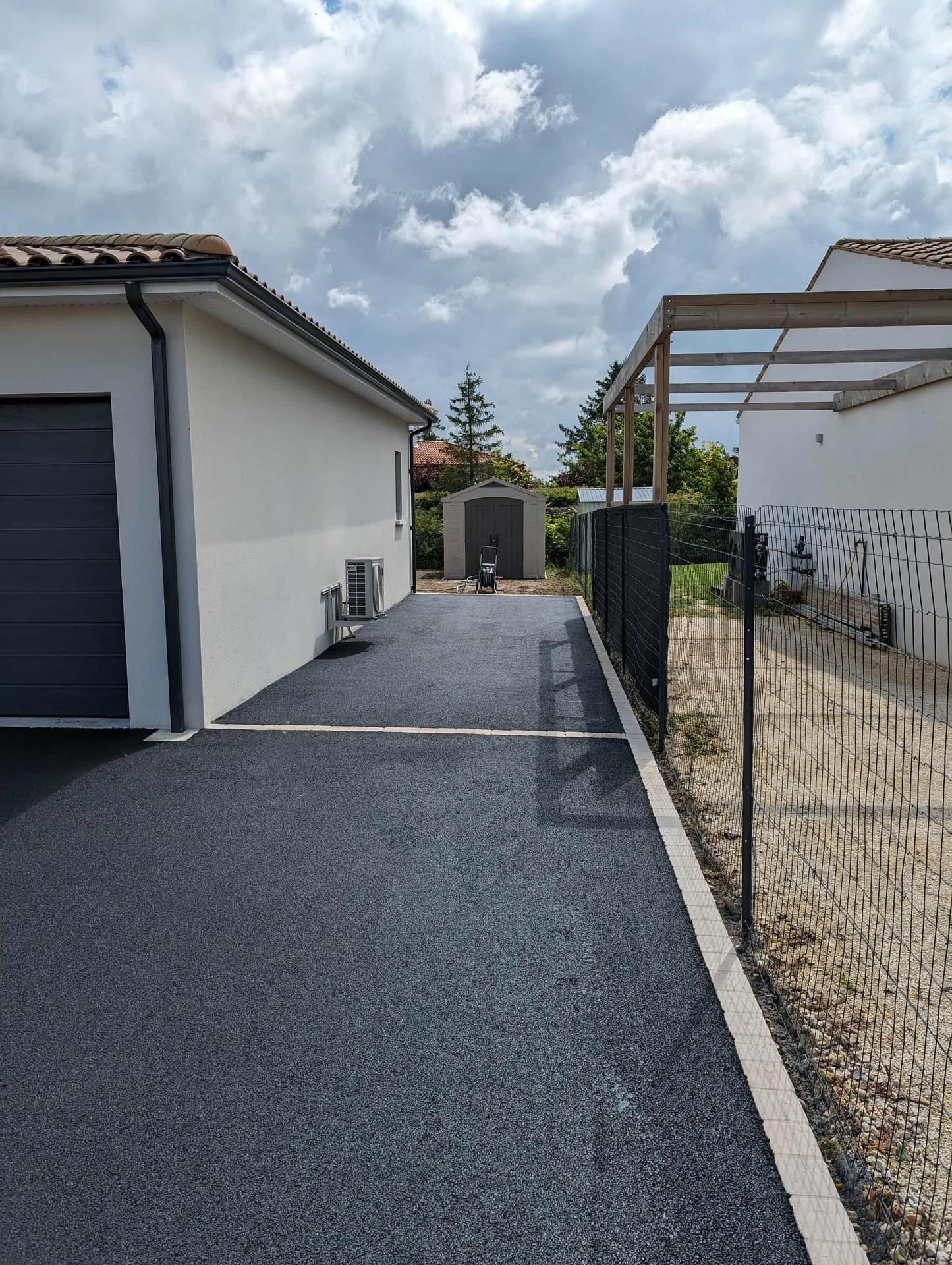 Allée asphaltée entre des bâtiments blancs avec une clôture métallique et un abri sous un ciel nuageux.