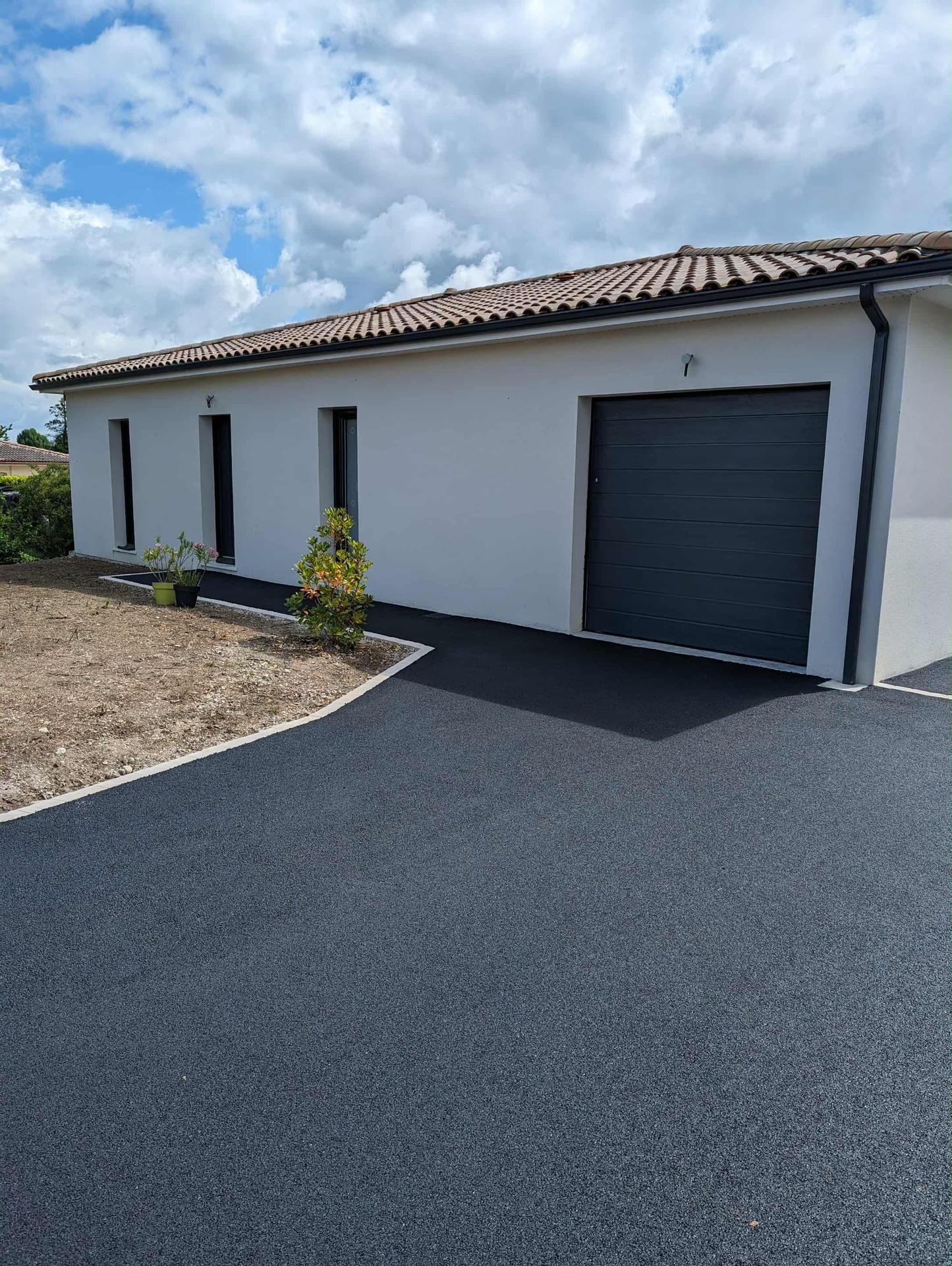 Maison blanche avec porte de garage gris foncé, allée asphaltée et toit de tuiles rouges sous un ciel nuageux.
