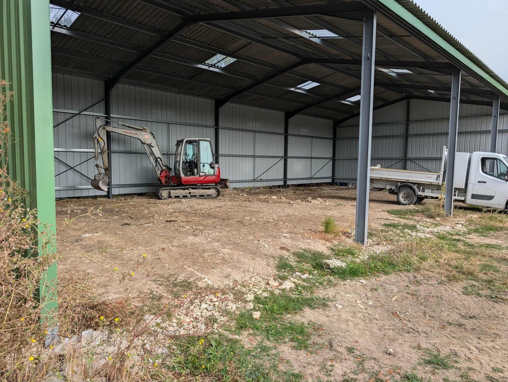 À l'intérieur d'un hangar métallique, une petite excavatrice et un camion blanc sont posés sur un sol en terre battue.