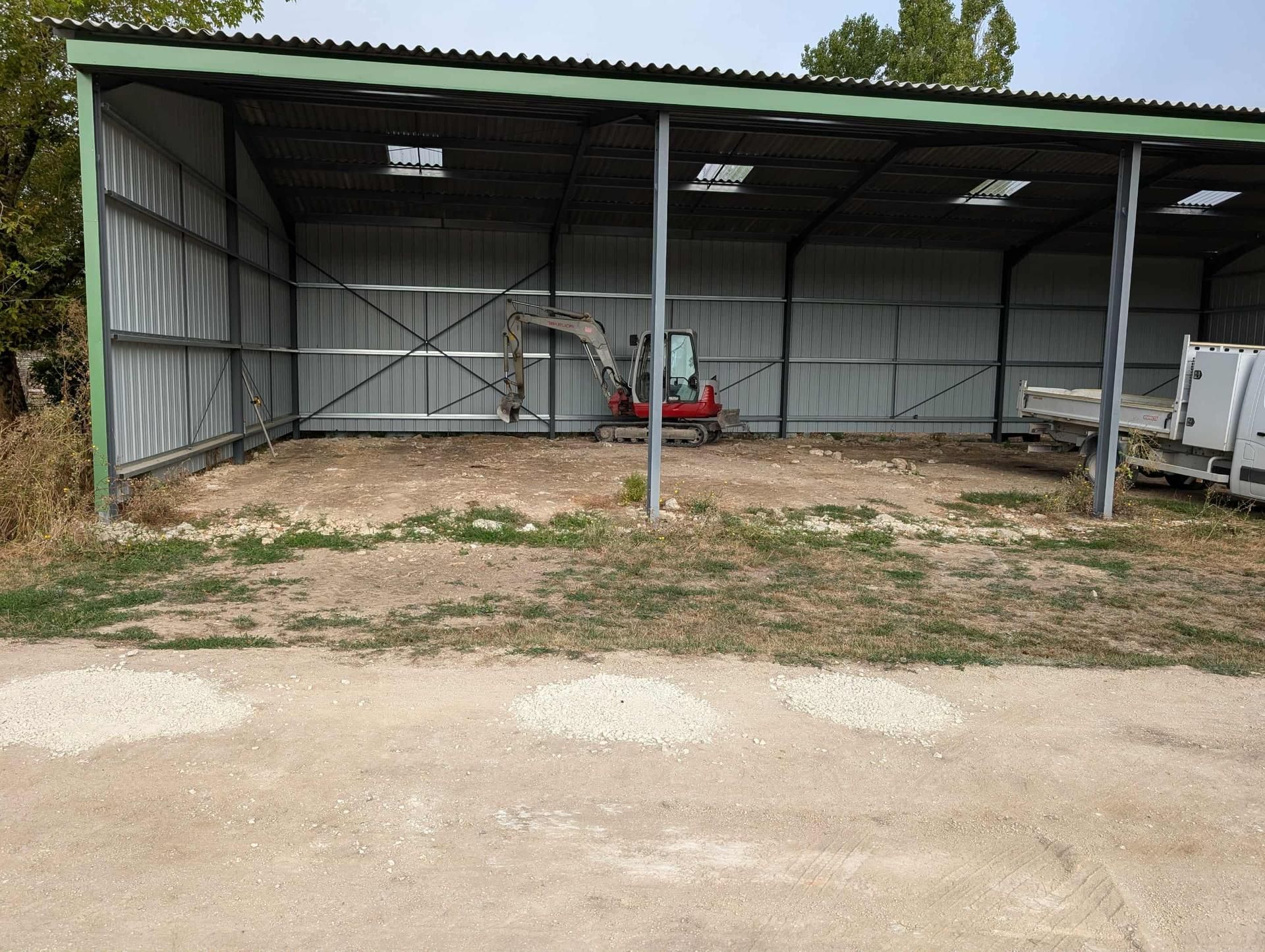 Mini-pelle à l'intérieur d'un hangar métallique ouvert sur les côtés. À l'extérieur, du gravier et quelques touffes de gazon.