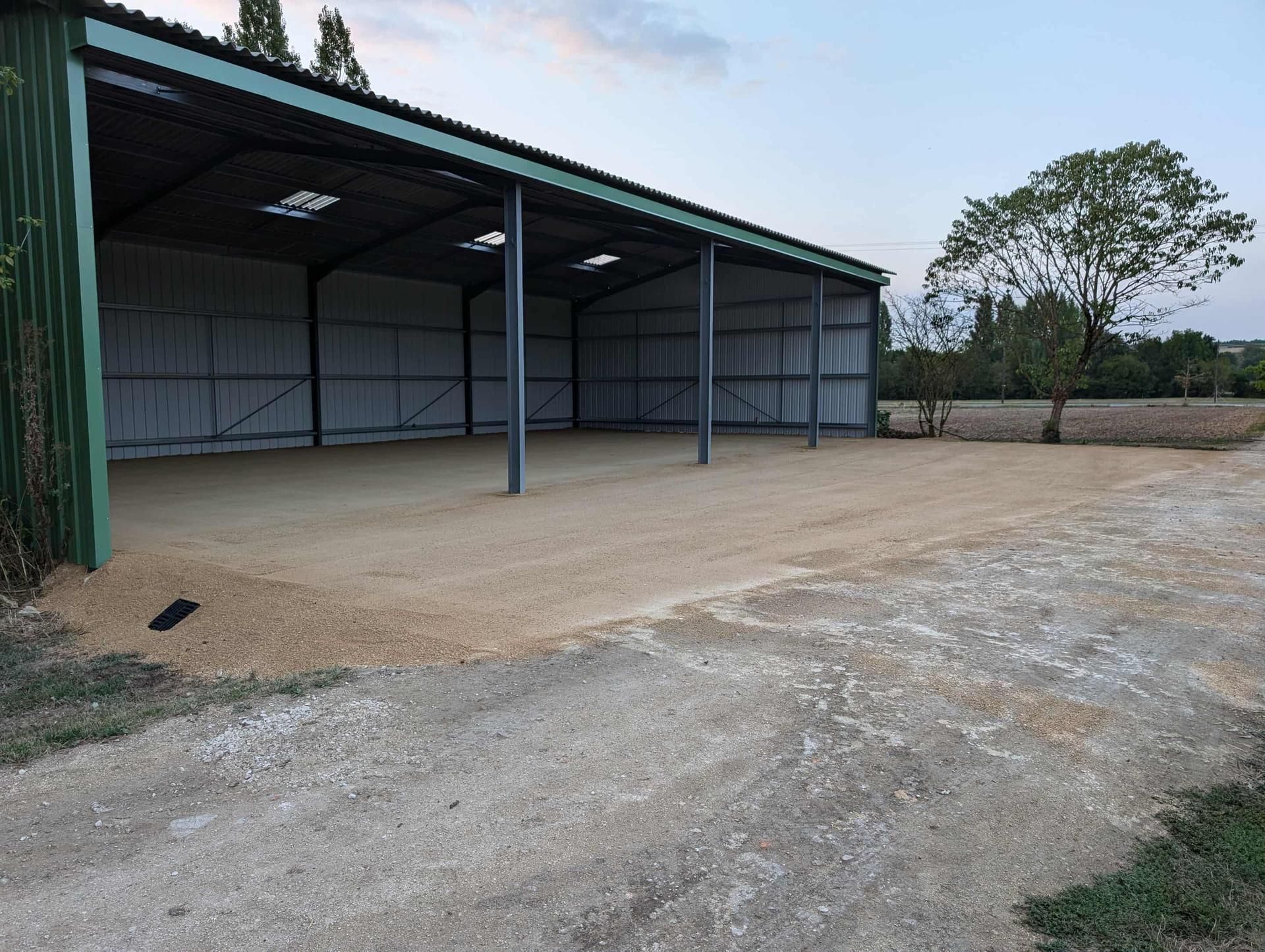 Abri métallique ouvert sur les côtés, avec un sol en gravier, situé en milieu rural avec un arbre et un chemin de terre.