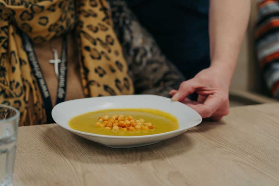 Eine Hand stellt eine weiße Schüssel mit Suppe, garniert mit Croutons, auf einen hellen Holztisch.