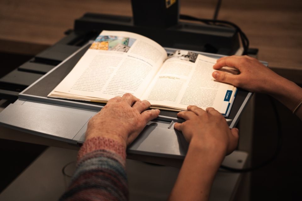 Zwei Personen legen ihre Hände auf ein offenes Buch, das unter einem Scanner positioniert ist, um das Lesen zu erleichtern.