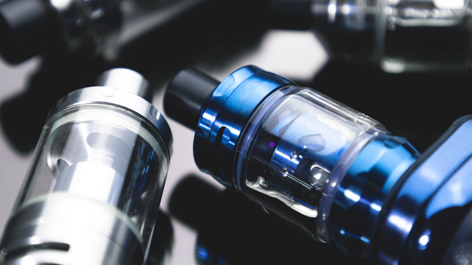 Gros plan de plusieurs stylos vape, un bleu et d'autres argentés, sur une surface noire réfléchissante.
