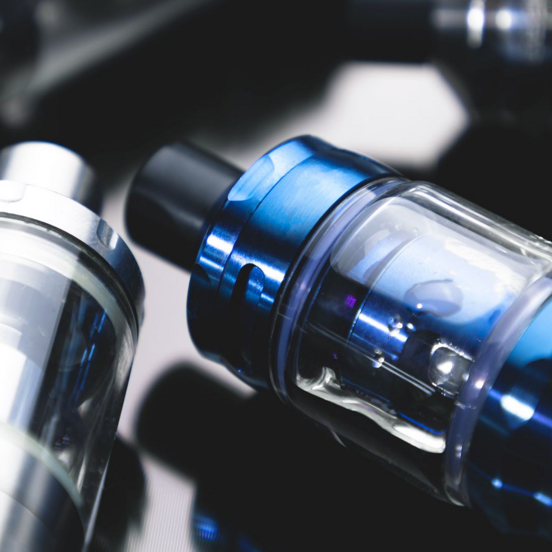 Gros plan de deux stylos vape, l'un argenté, l'autre bleu, sur une surface noire réfléchissante.