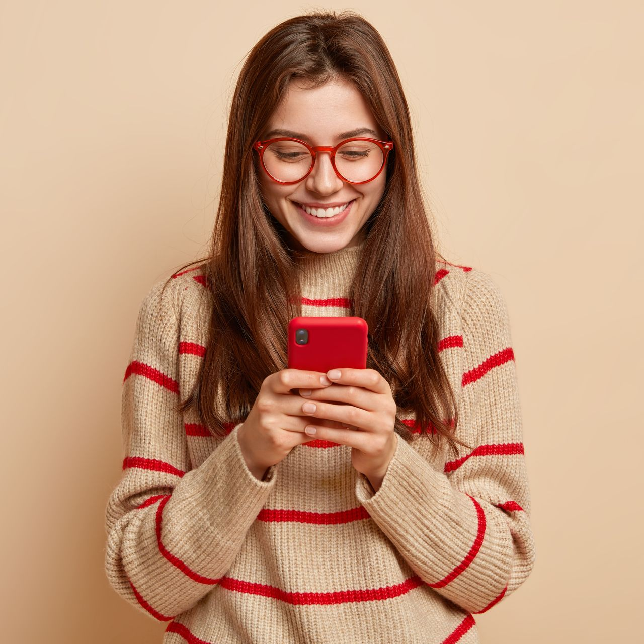 Une femme portant des lunettes et un téléphone rouge sourit en regardant l'écran, vêtue d'un pull rayé.