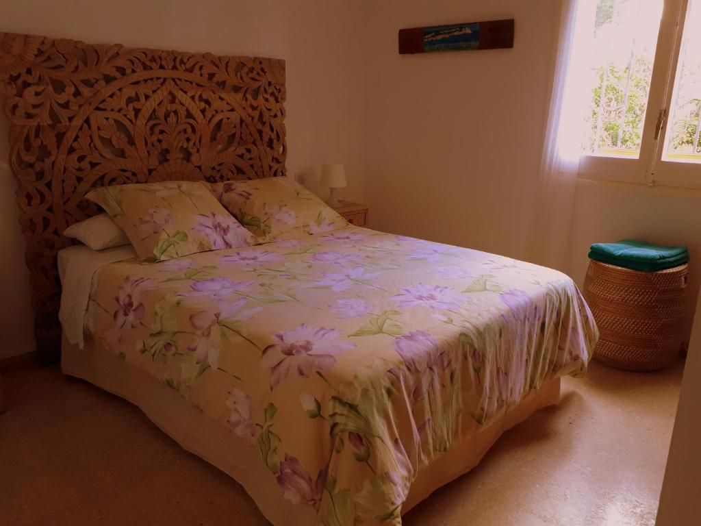 Cama impecablemente hecha con ropa de cama floral en un dormitorio soleado con cabecero de madera tallada y cesta de mimbre.
