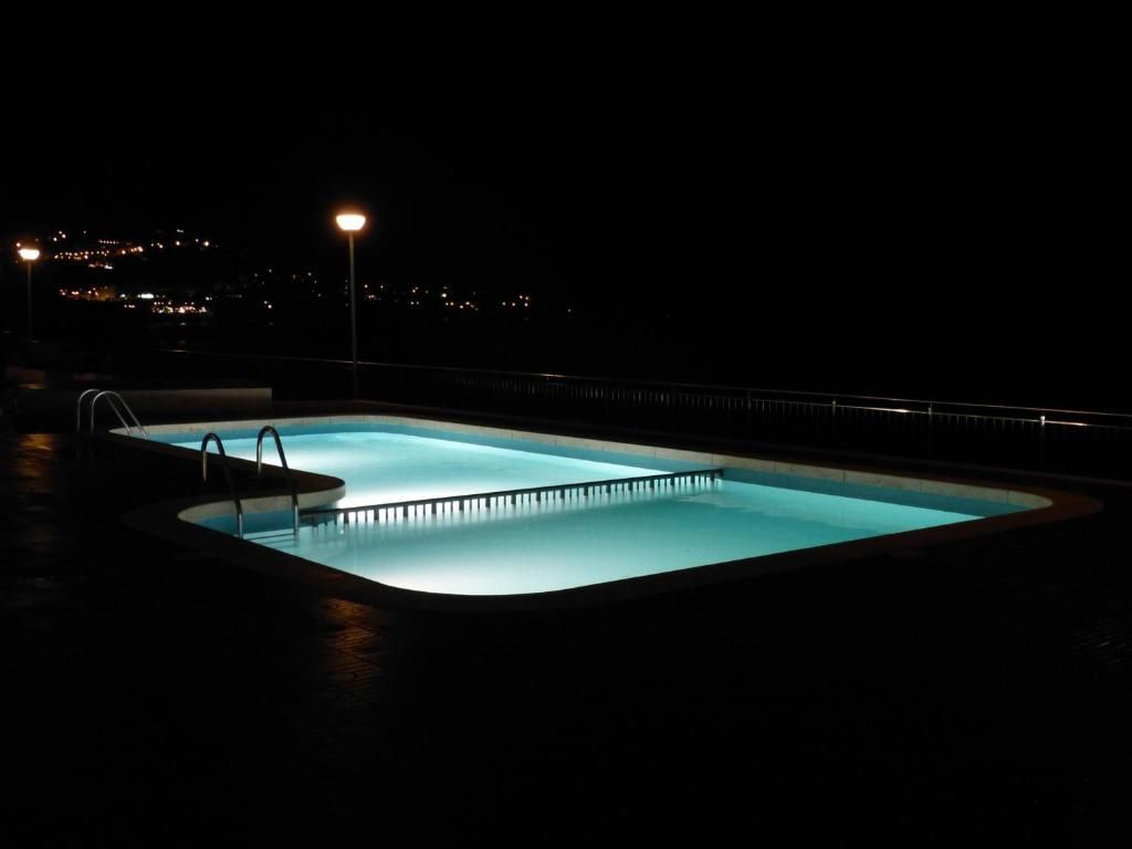 Piscina exterior nocturna que brilla en azul junto a una terraza oscura y las luces de la ciudad a lo lejos.