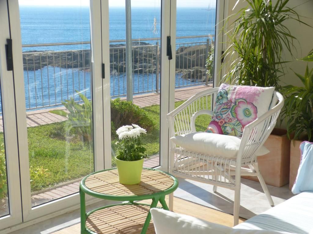 Acogedora terraza acristalada con silla de mimbre, mesita, maceta con flores y vistas al océano.