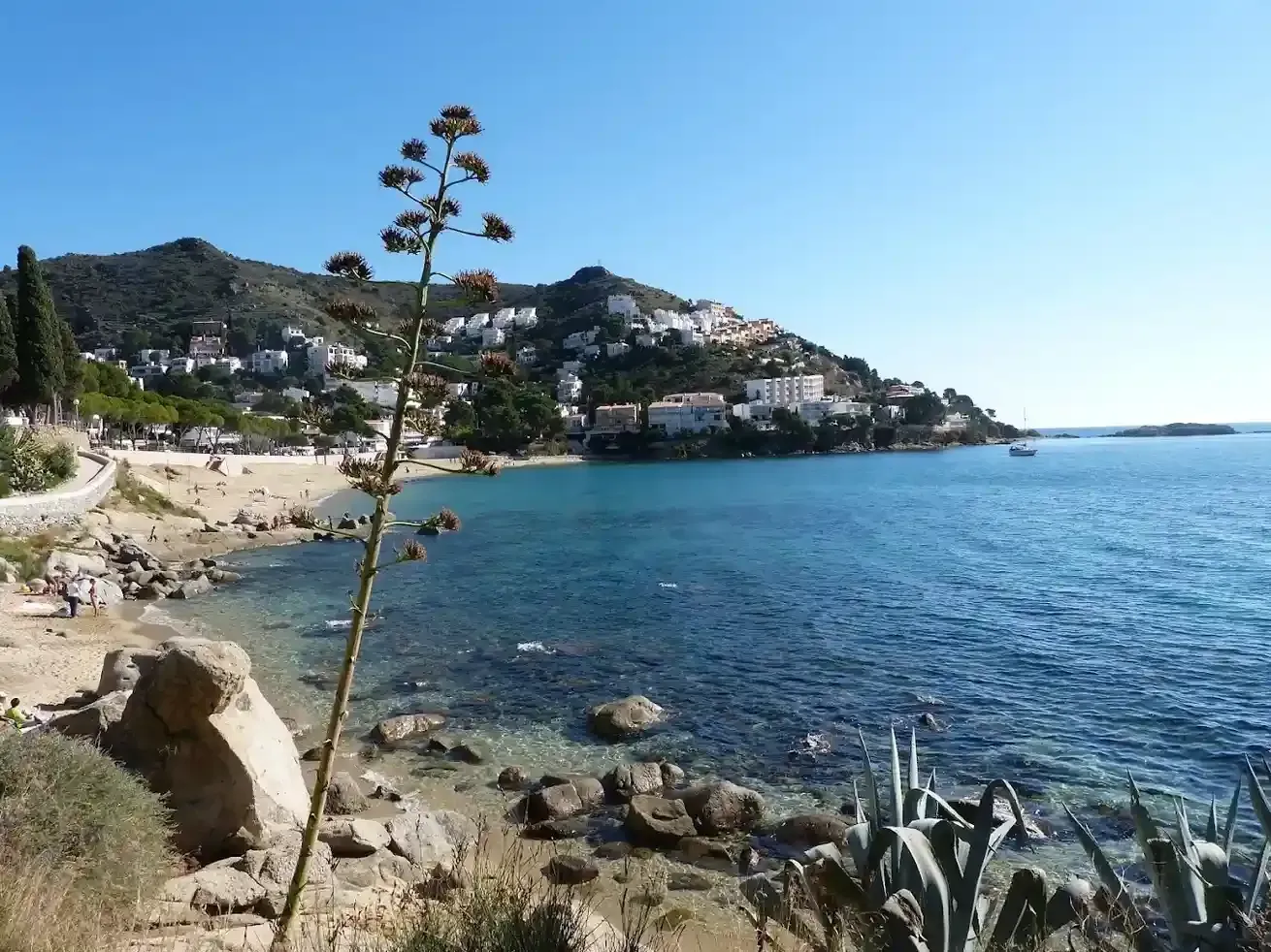 Playa rocosa junto a una bahía azul, con casas en la ladera y un cielo despejado.