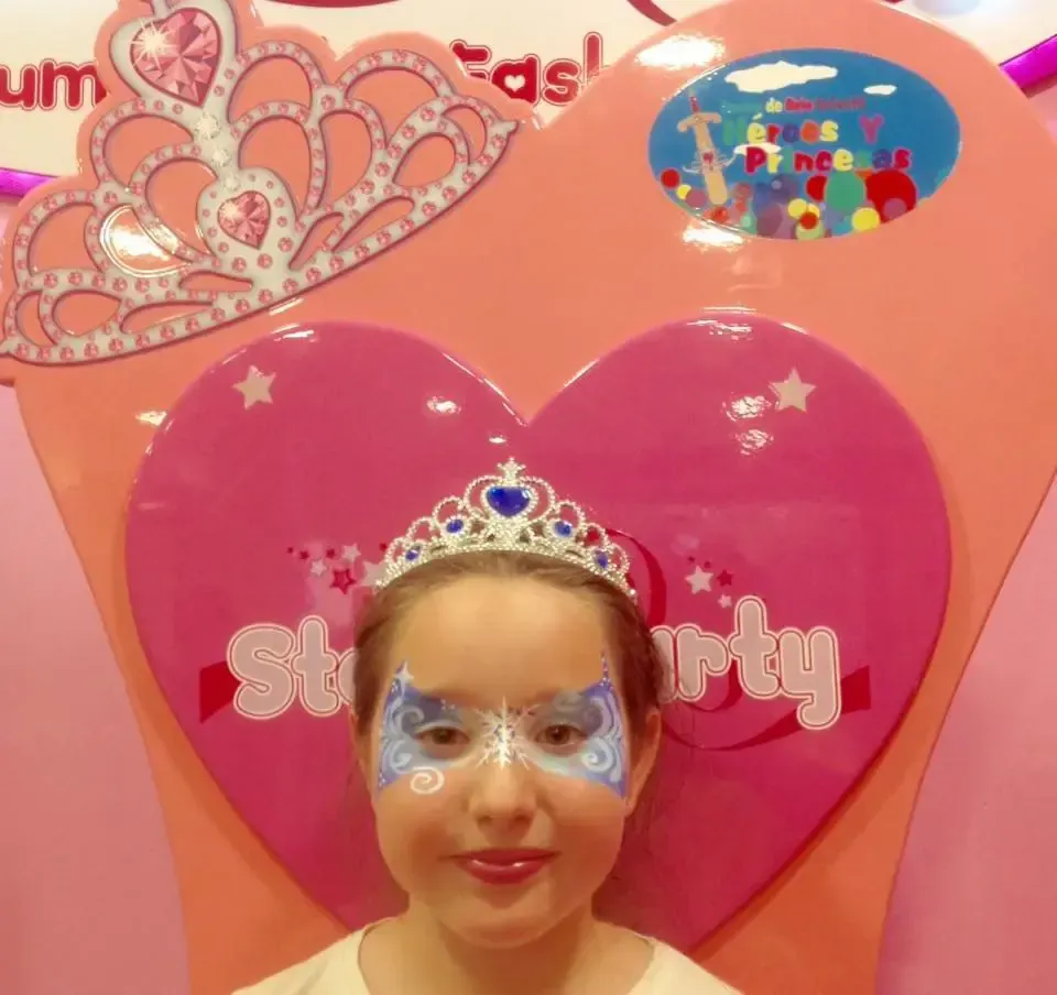 Niña con pintura facial y tiara frente a un fondo de corazones rosas que anuncia una 