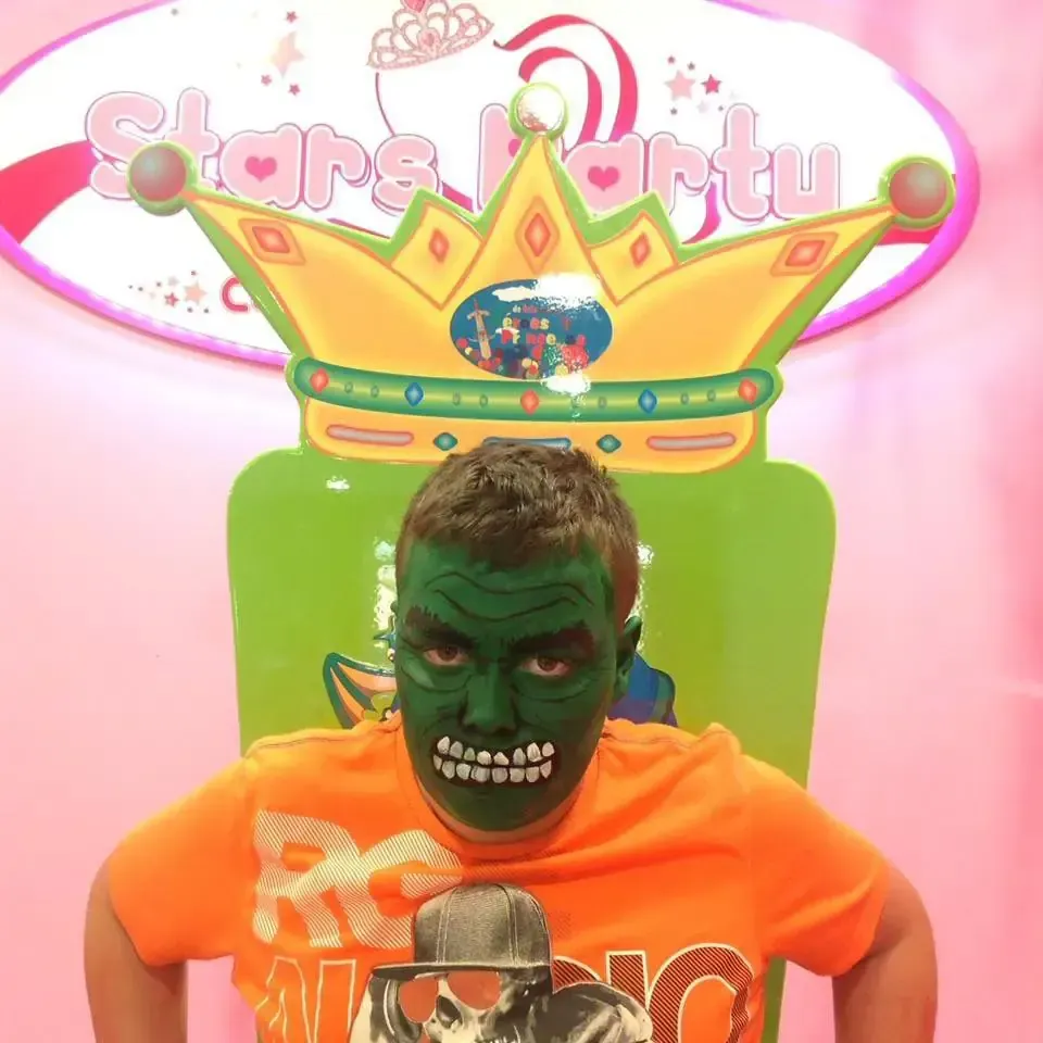 Persona con la cara pintada de verde, sentada en una silla trono verde, frente a un fondo de 