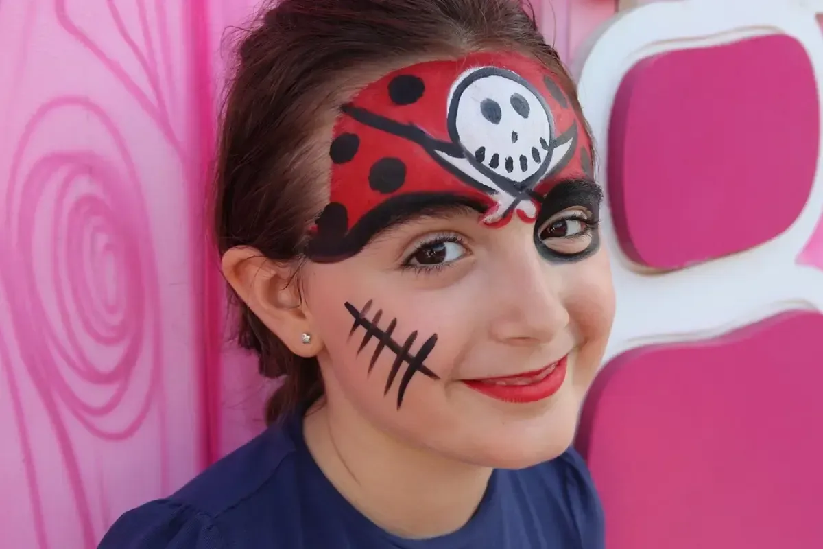Chica con cara de pirata pintada en rojo y negro, sonriendo. Fondo rosa.