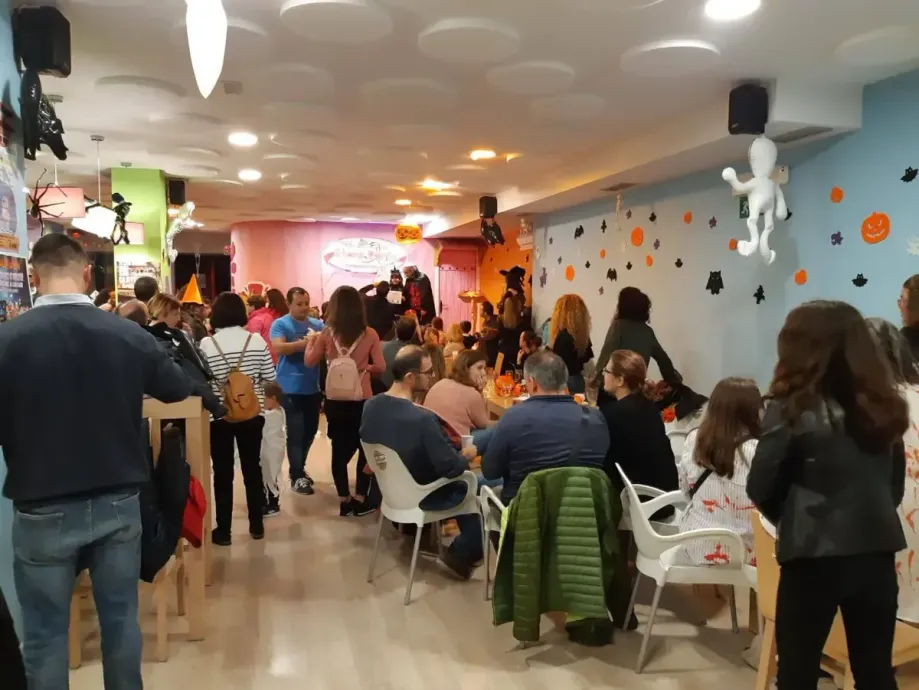 Un restaurante lleno de gente, sentada y de pie. Mesas y paredes decoradas; iluminación cálida.