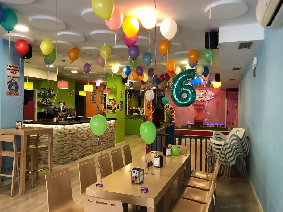 Interior de un salón de fiestas con globos. Una mesa está preparada para los invitados. Decoración en verde, rosa y amarillo.