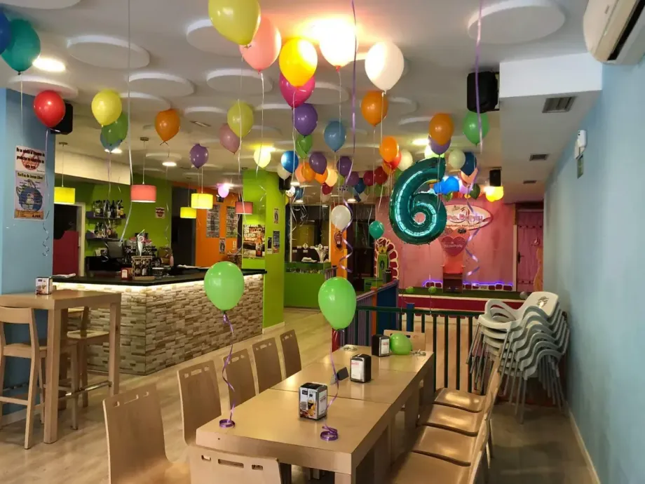 Interior de un salón de fiestas con globos. Una mesa está preparada para los invitados. Decoración en verde, rosa y amarillo.