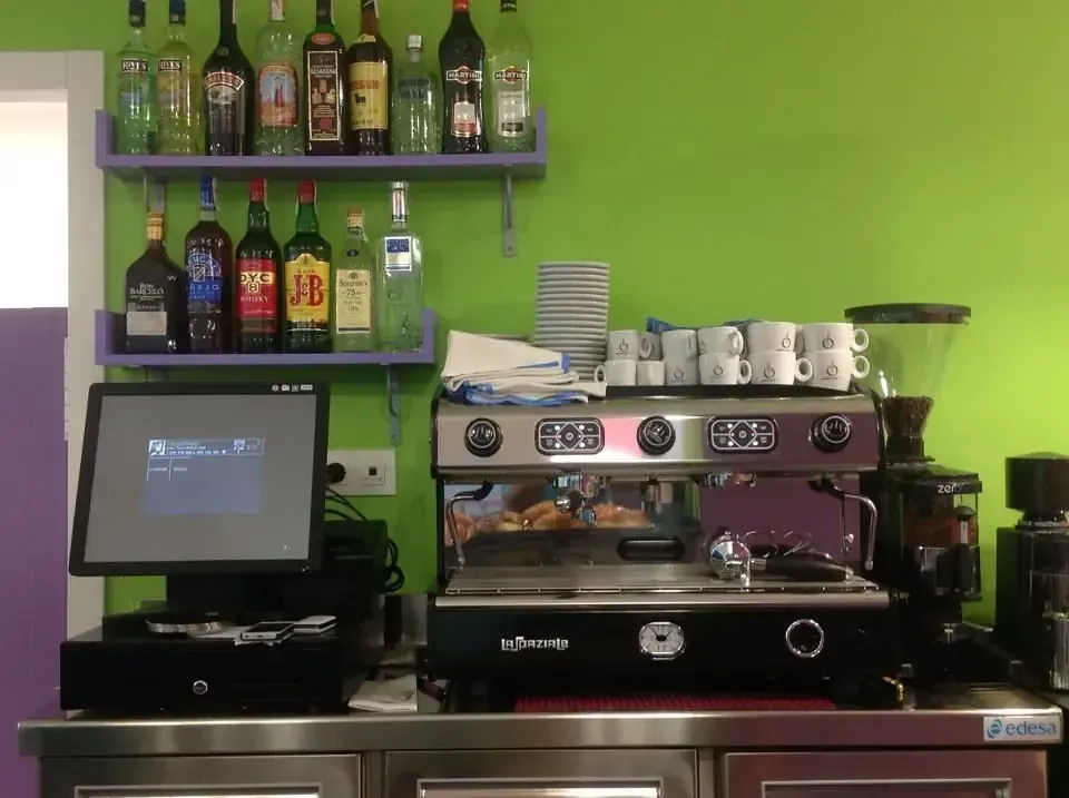 Máquina de café y sistema de punto de venta sobre mostrador de acero inoxidable contra una pared verde con estantes de botellas.