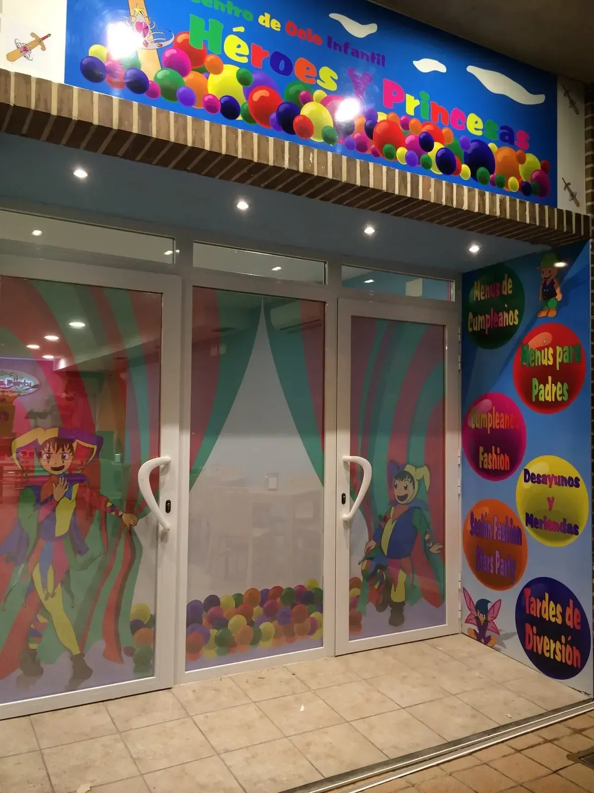 Entrada a un área de juegos infantiles con coloridas decoraciones de payasos, piscina de pelotas visible a través de las puertas y un cartel azul.