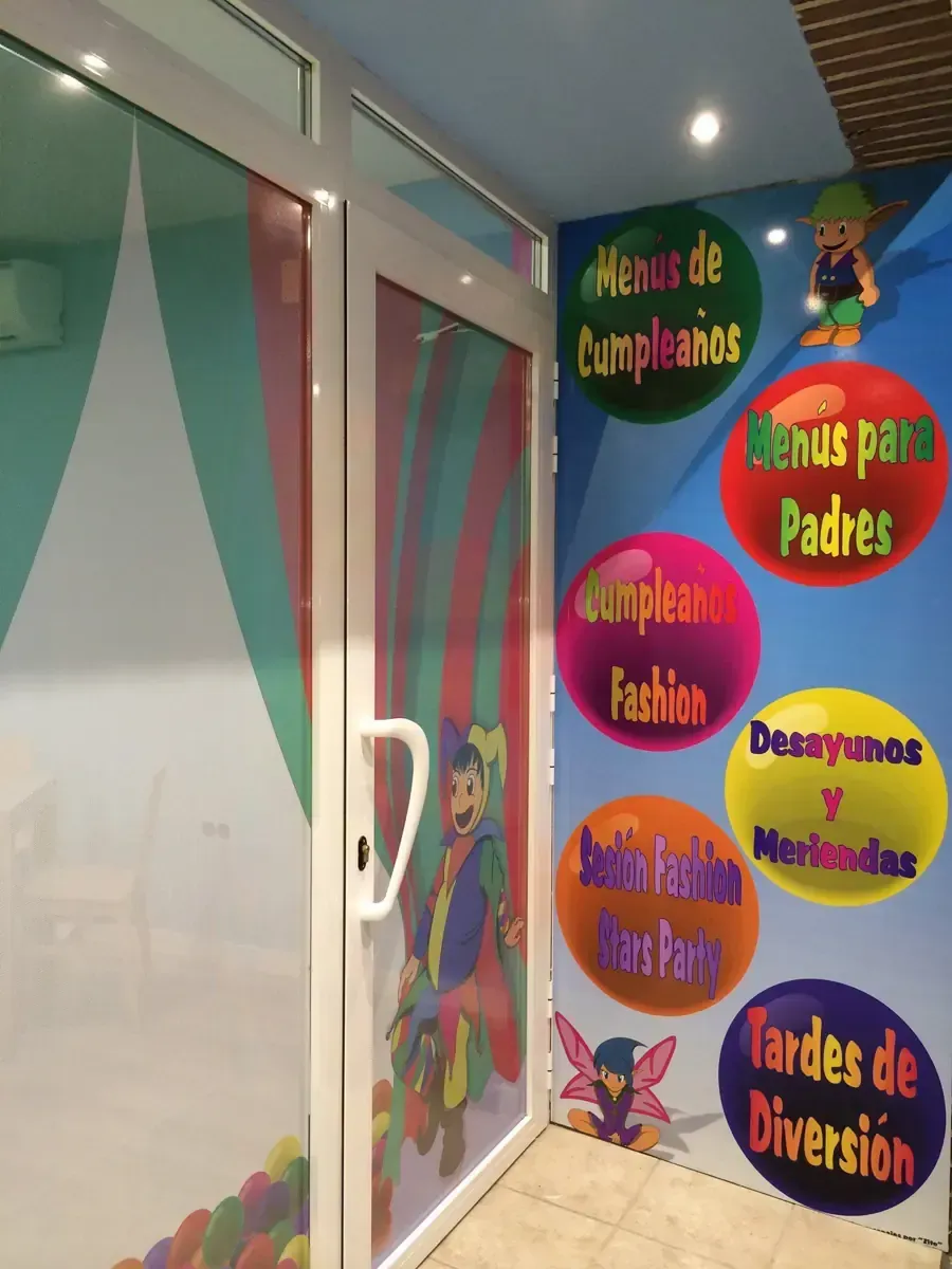 Entrada a un salón de fiestas infantiles. Puerta con un personaje de dibujos animados. Letreros coloridos en la pared: menús de cumpleaños, menús para padres.
