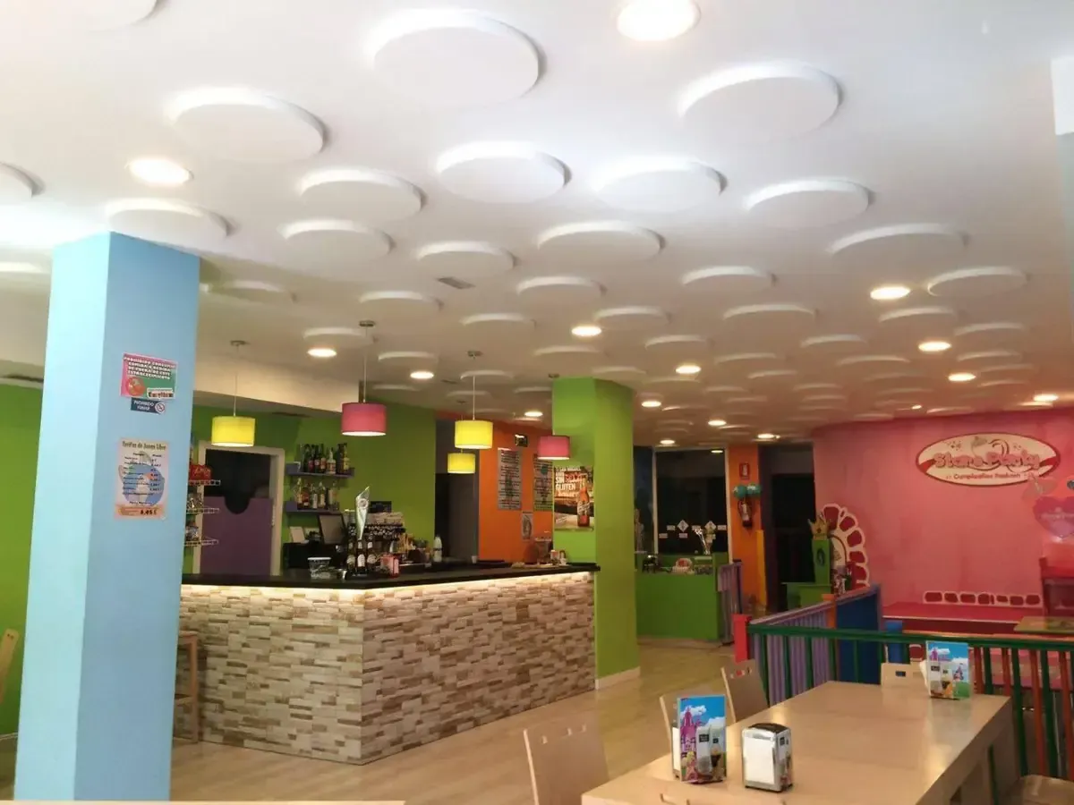 Interior de cafetería colorido con barra y techo circular. Paredes, mesas e iluminación en tonos rosa, azul y verde.