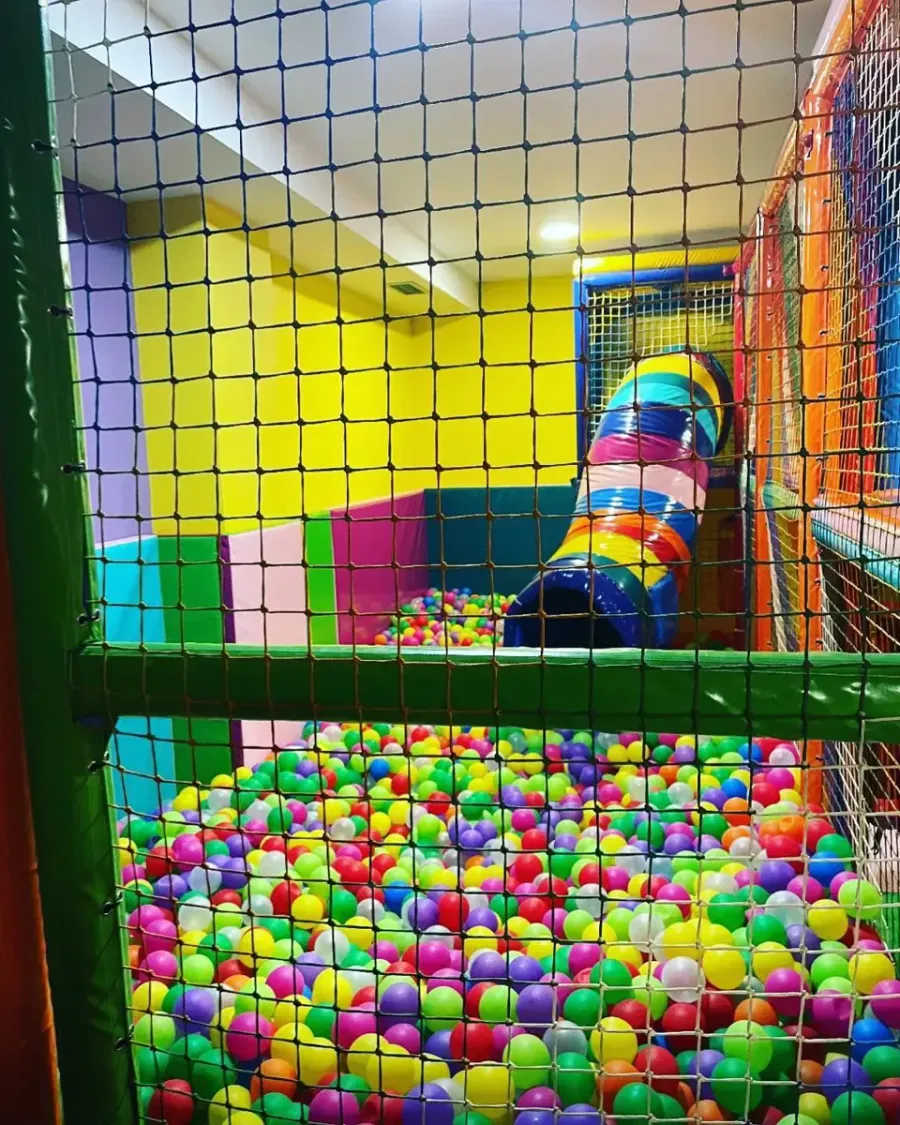 Piscina de bolas con pelotas de colores, tobogán tipo túnel y red de seguridad. Se ven paredes amarillas, verdes, azules y rosas.