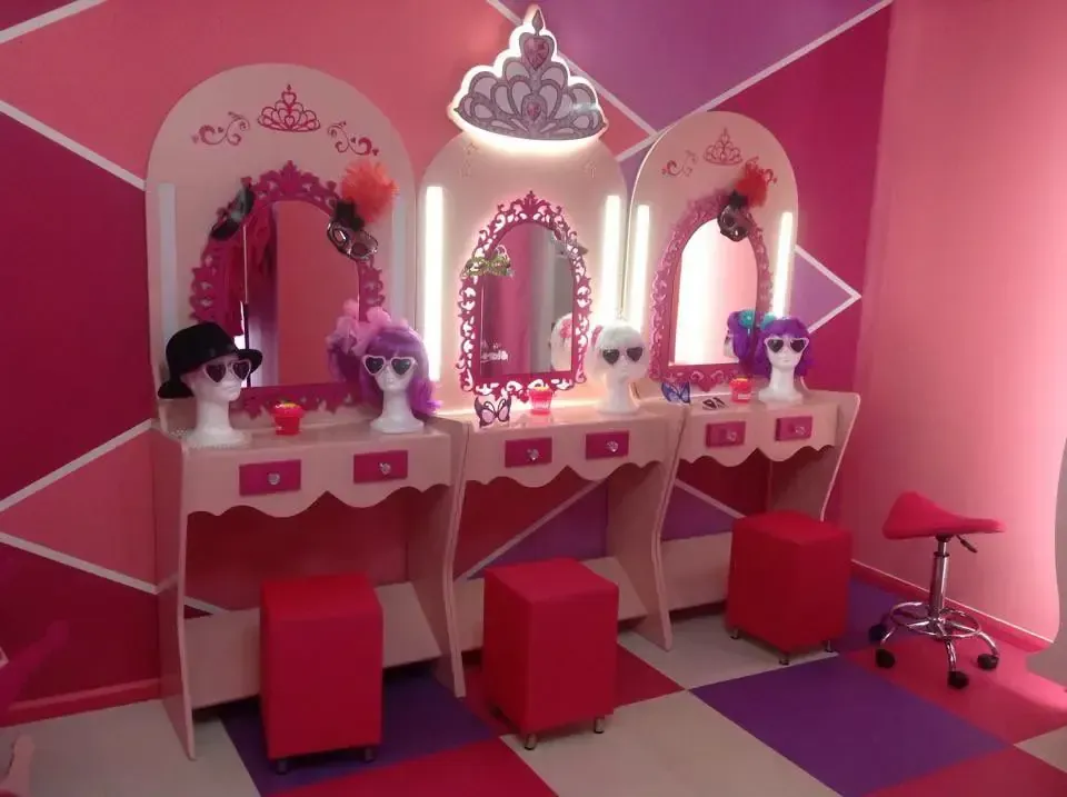 Tocador rosa con tres estaciones, pelucas y taburetes. Decoración temática con lámpara de tiara y suelo estampado.