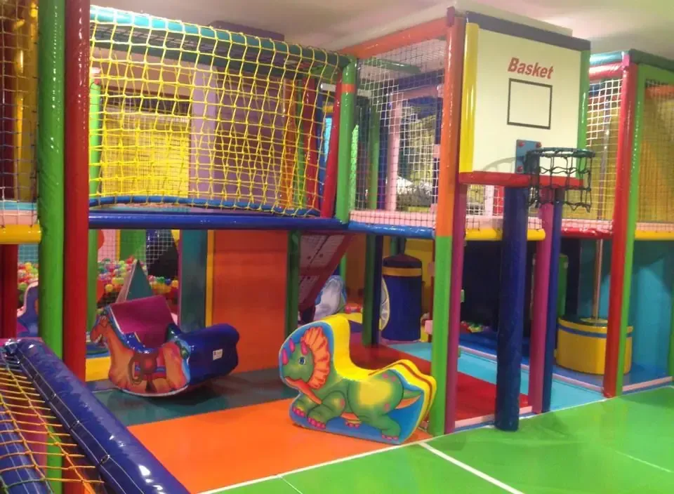 Área de juegos interior con coloridas estructuras para escalar, aro de baloncesto y juguetes mecedores.