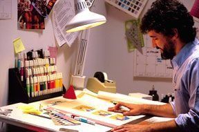 Artista en una mesa de dibujo, iluminada por una lámpara, trabajando en ilustraciones; marcadores y muestras de color visibles.