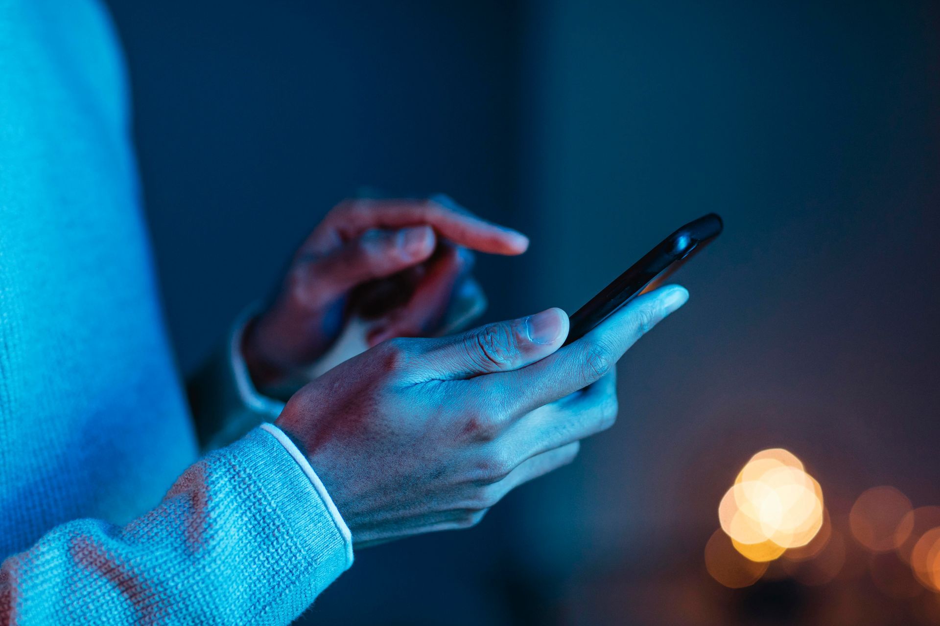 Manos sosteniendo e interactuando con un teléfono inteligente, iluminadas por luz ambiental azul y naranja.