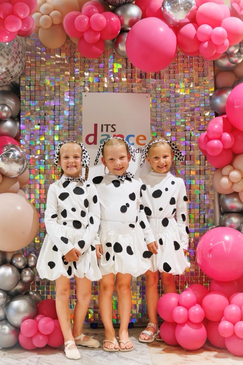 Three little girls in dalmatian dresses are standing in front of a wall of balloons.