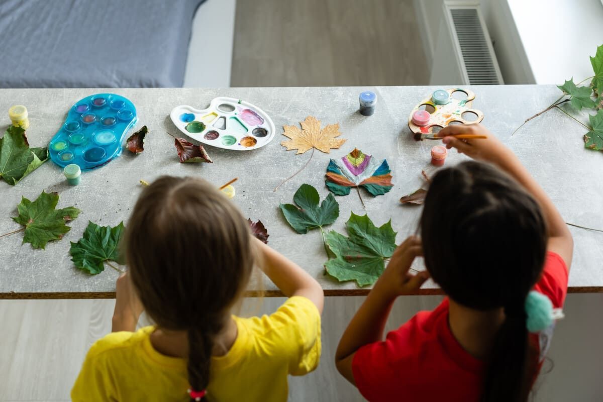 Dos niños pintan hojas de otoño en una mesa. Se ven paletas y pinceles.