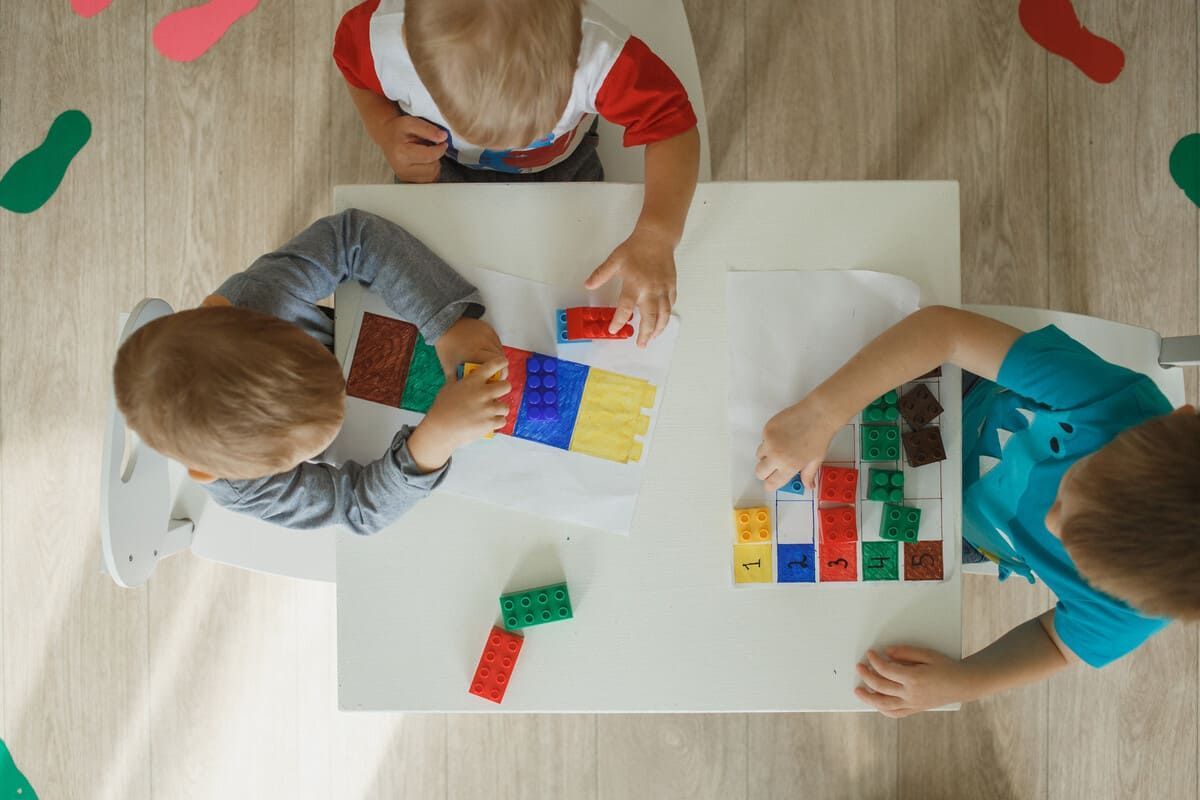 Tres niños pequeños construyen con bloques de colores en una mesa blanca.
