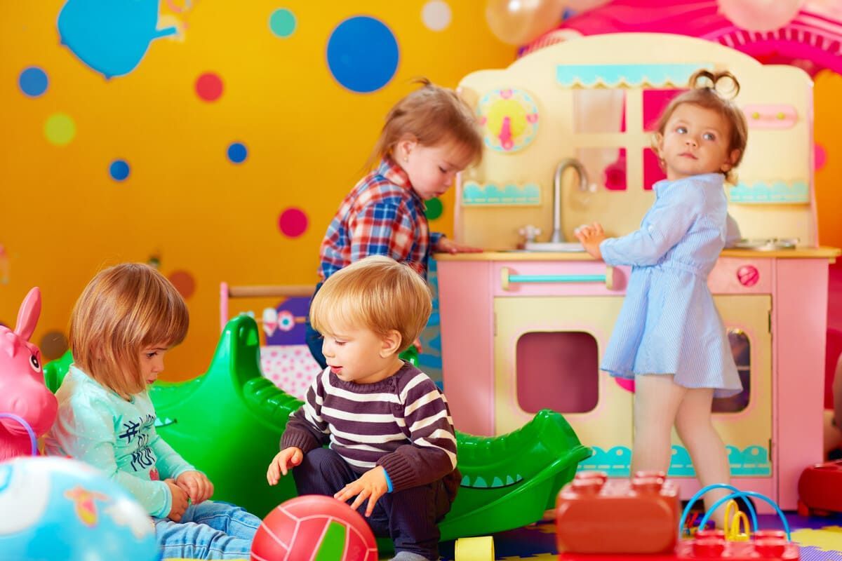 Niños jugando en una sala de juegos colorida con una cocina de juguete, juguetes mecedores y una pelota.