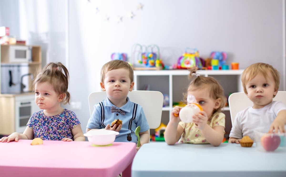 Cuatro niños pequeños en una mesa en una guardería, algunos comiendo, con juguetes coloridos en el fondo.