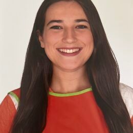 Mujer con cabello largo y oscuro, sonriente, vistiendo una blusa roja y blanca con un borde verde, sobre un fondo blanco.
