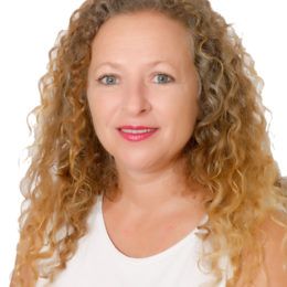 Mujer con cabello rubio rizado sonriendo, vistiendo un top blanco, frente a un fondo blanco.