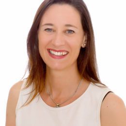Mujer de cabello castaño sonriendo, vistiendo un top blanco y un collar de plata.