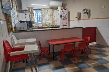 Comedor infantil con sillas rojas y una mesa. Tronas y utensilios de cocina a la vista.